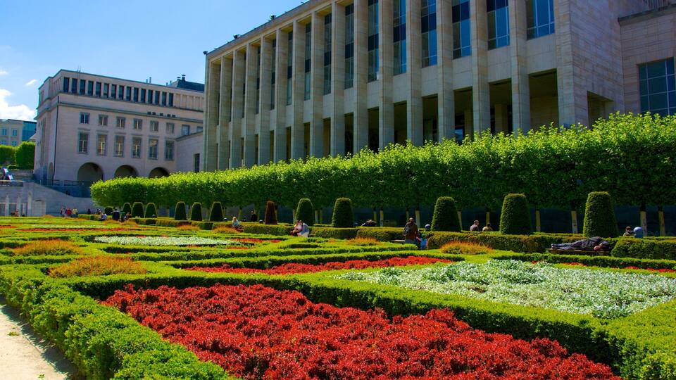 Um jardim formal com sebes geometricamente modeladas e flores vermelhas, em frente a um grande edifício com colunas.