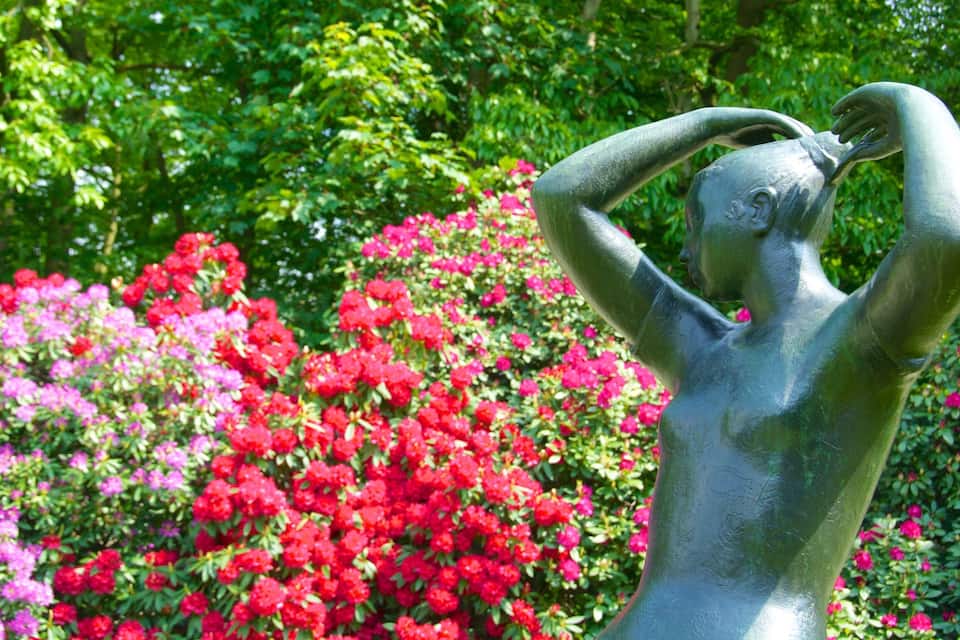 Een bronzen beeld van een vrouw met opgetrokken armen in een tuin vol rode en roze bloemen.