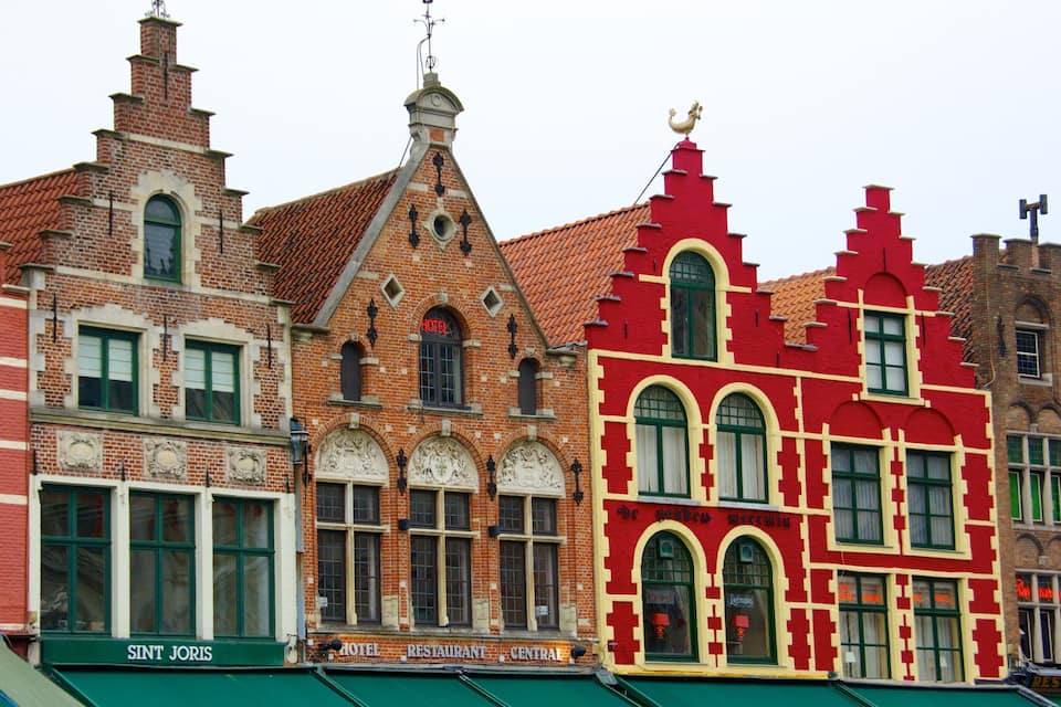 Uma fileira de edifícios coloridos com estilos arquitetônicos distintos, incluindo um prédio vermelho com janelas verdes e um prédio de tijolos com telhado pontiagudo.