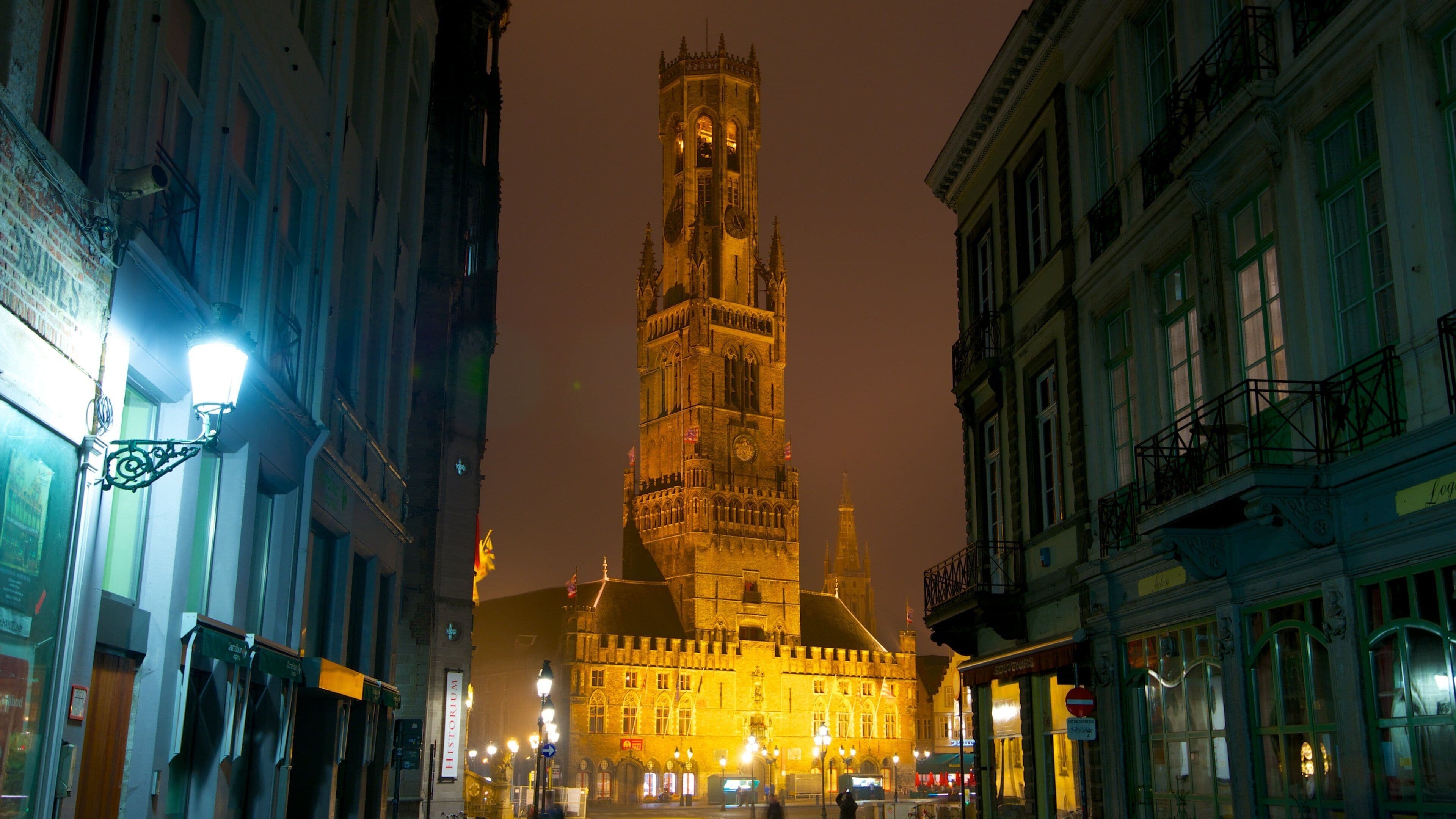 Uma torre histórica com relógio, rodeada por edifícios iluminados à noite.