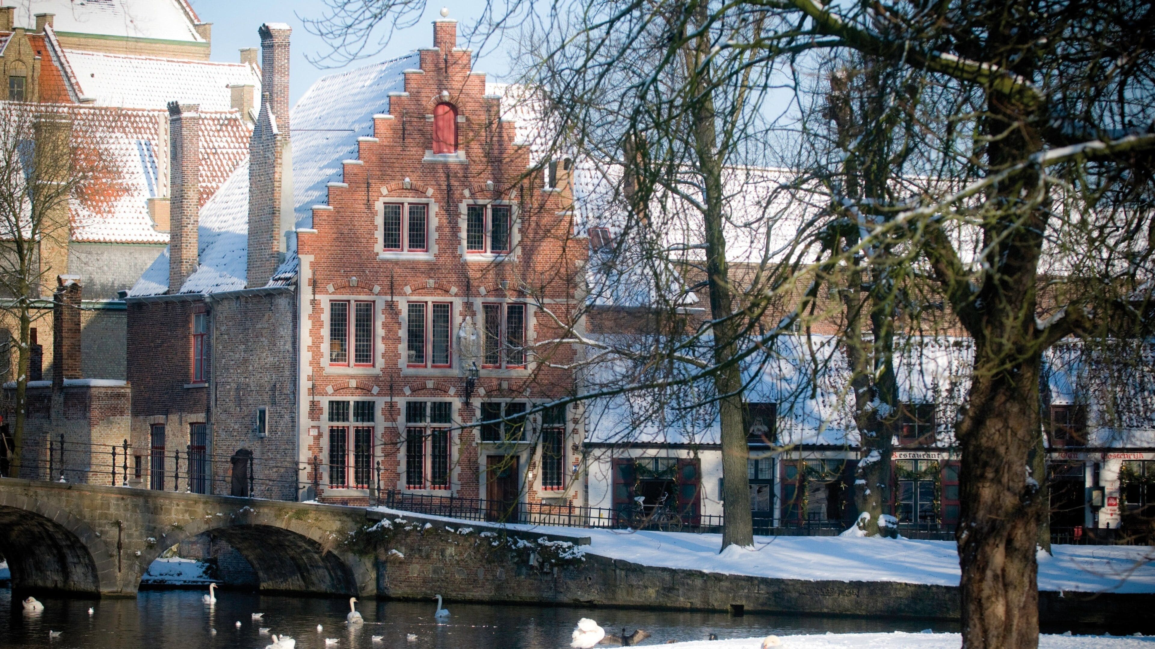 A snow-covered canal with swans, a stone bridge, and historic brick buildings.