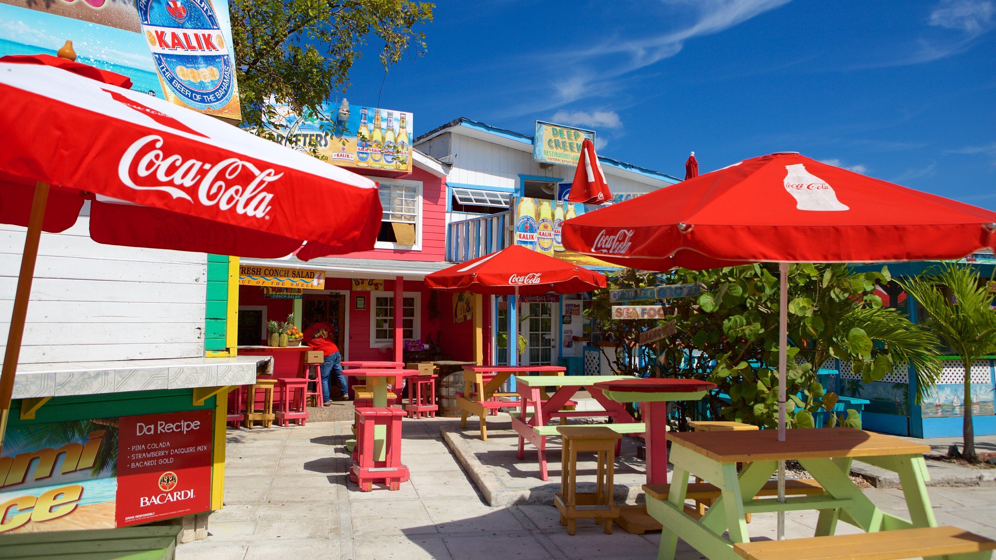 Un área de comedor al aire libre con coloridas sombrillas rojas de Coca-Cola, un letrero que dice 'Ensalada Fresca de Caracol' y un letrero 'Da Recipe' que enumera Piña Colada, Batido de Fresa y Bacardi Gold.