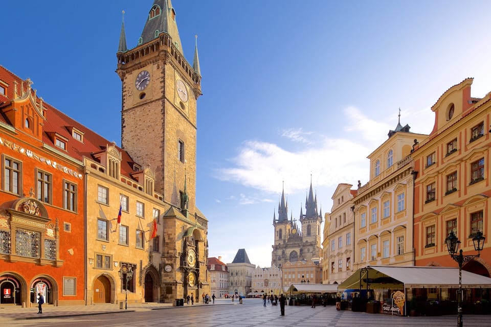 A historic square with a prominent clock tower, traditional European architecture, and a clear blue sky.