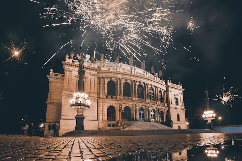 Um edifício histórico com fachada imponente, uma grande fonte em frente e fogos de artifício explodindo no céu.
