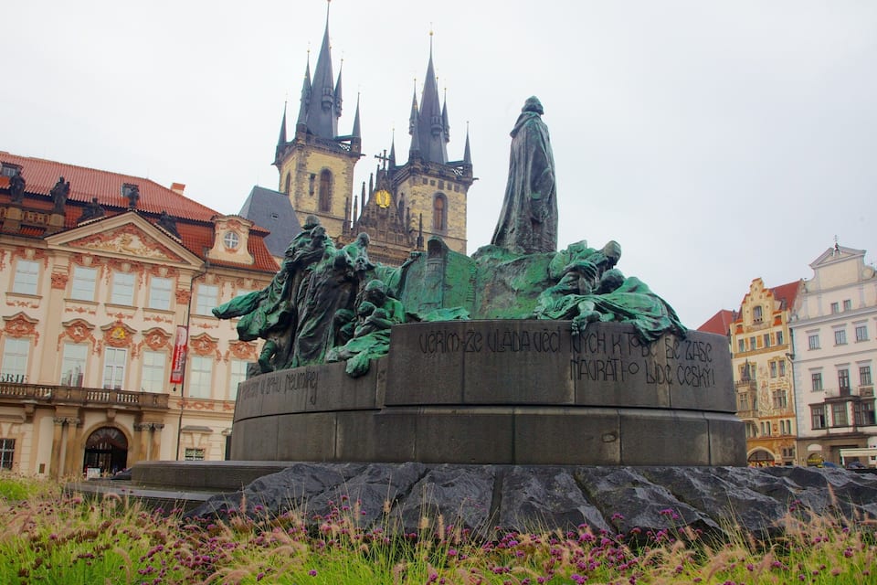Uma estátua retratando uma cena de batalha, com inscrições, ao fundo edifícios históricos e o topo de uma igreja.