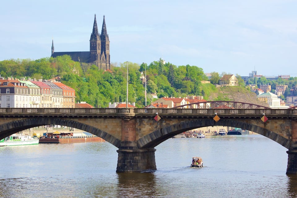 A stone bridge over a river with a boat in the foreground and a cathedral in the background.