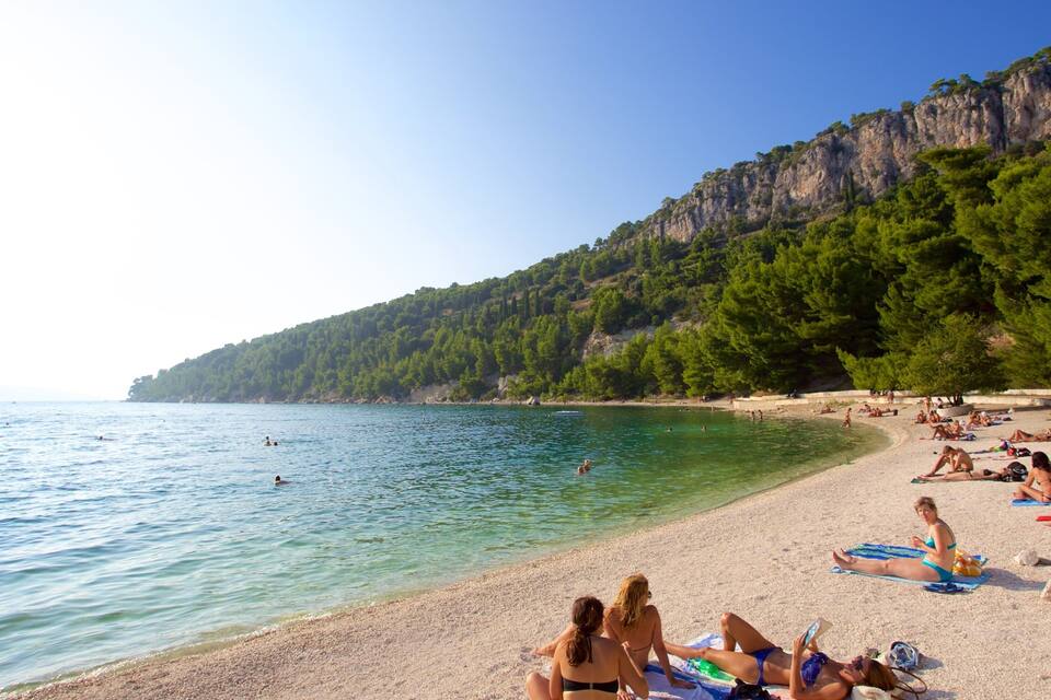 A sunny beach with clear turquoise water, people sunbathing, and a rocky cliff with trees.