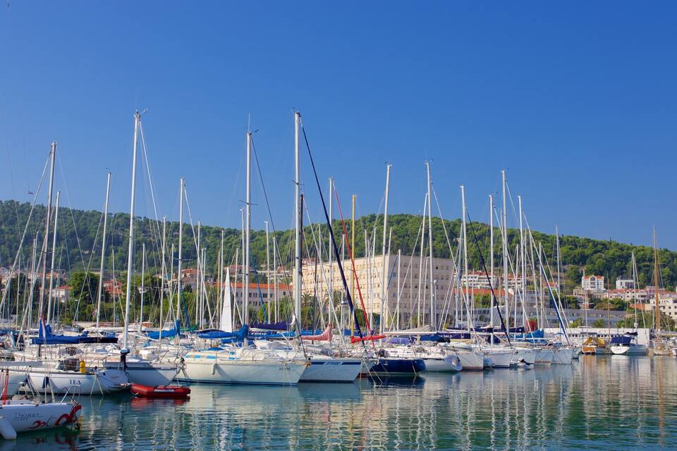 Una marina con numerosi aliscafi ormeggiati, un cielo azzurro limpido e una collina con edifici sullo sfondo.