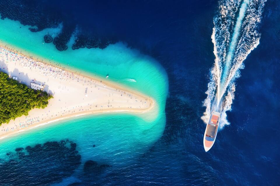 An aerial view of a beach with clear turquoise waters, a sandy shore, and a boat creating a wake in the ocean.