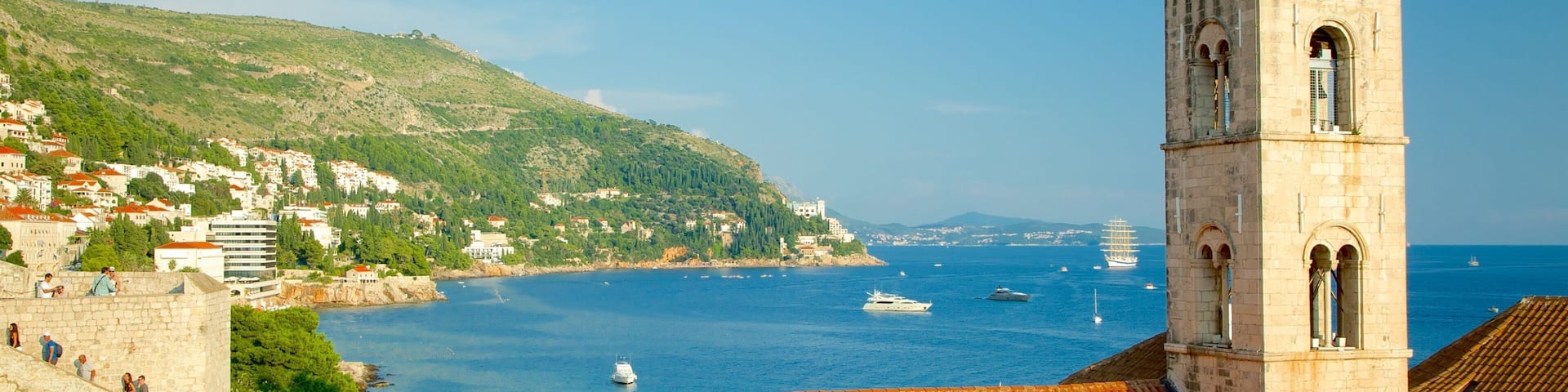 A coastal town with a prominent tower, terracotta rooftops, and a clear blue sea with boats.
