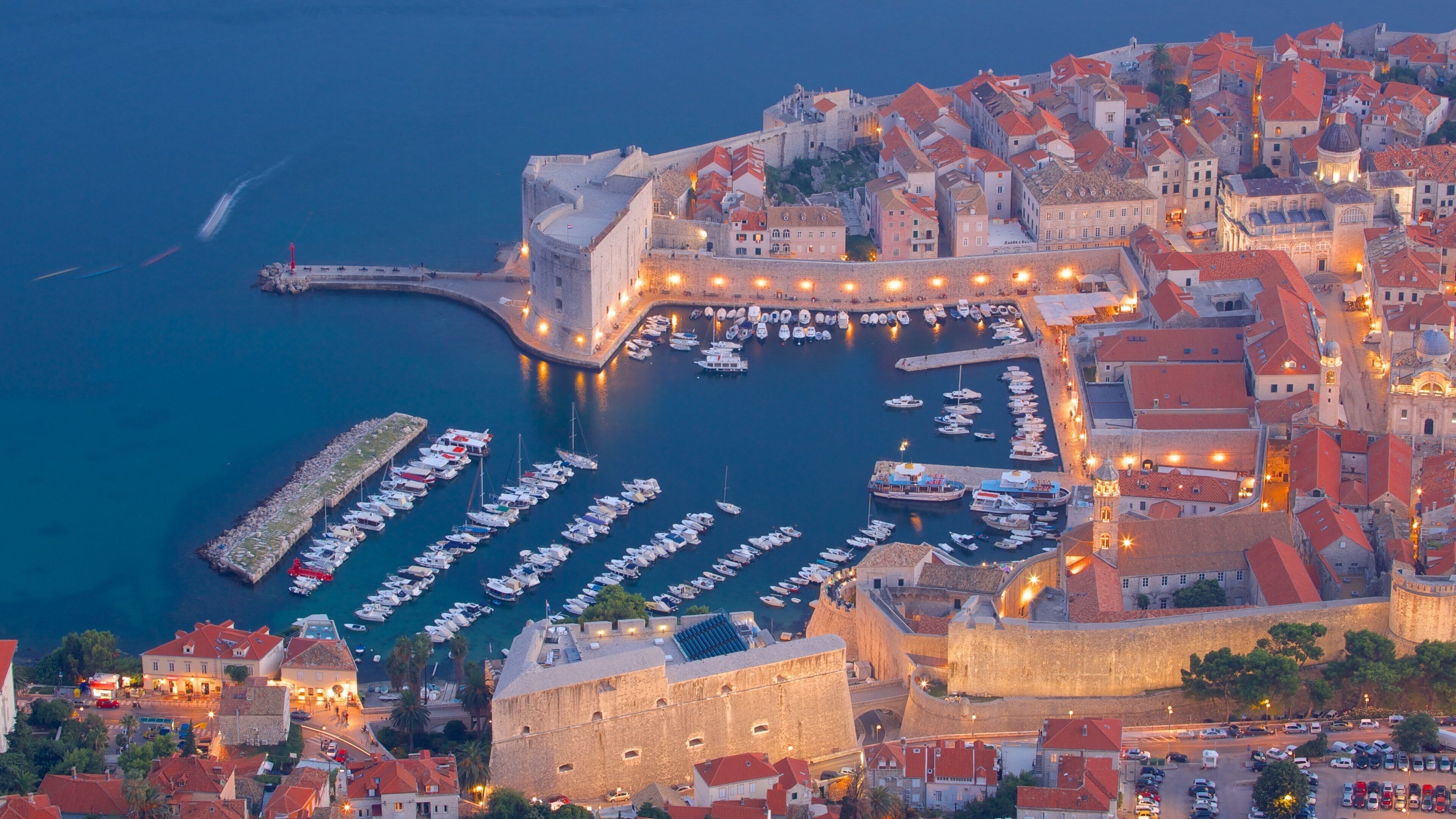 A coastal city with a historic fort, a harbor filled with boats, and a densely built urban area with red-roofed buildings.