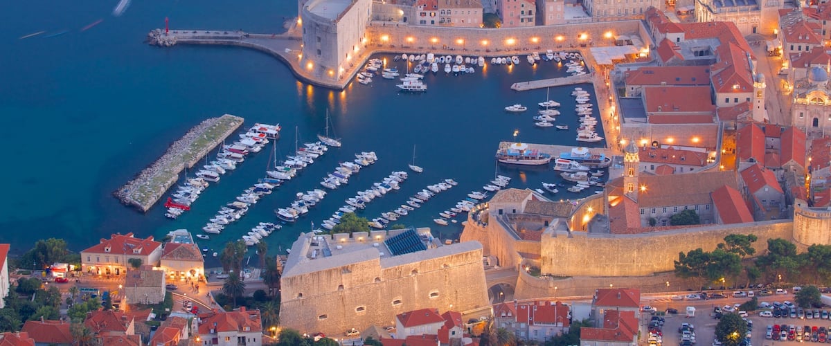 A coastal city with a historic fort, a harbor filled with boats, and a densely built urban area with red-roofed buildings.