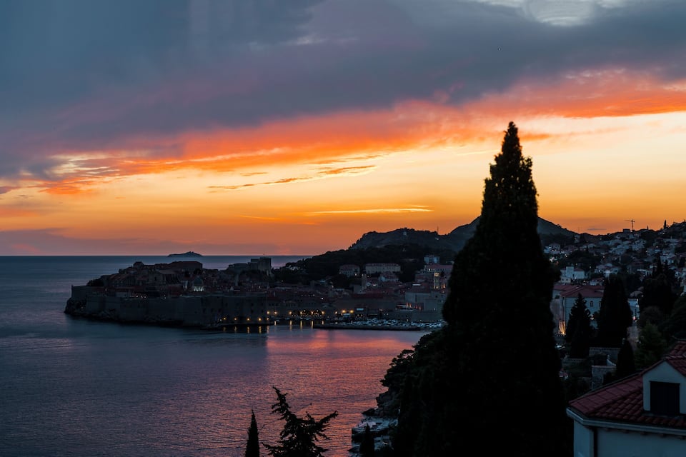 Una città costiera al tramonto con una torre imponente, mare calmo e una penisola con edifici.