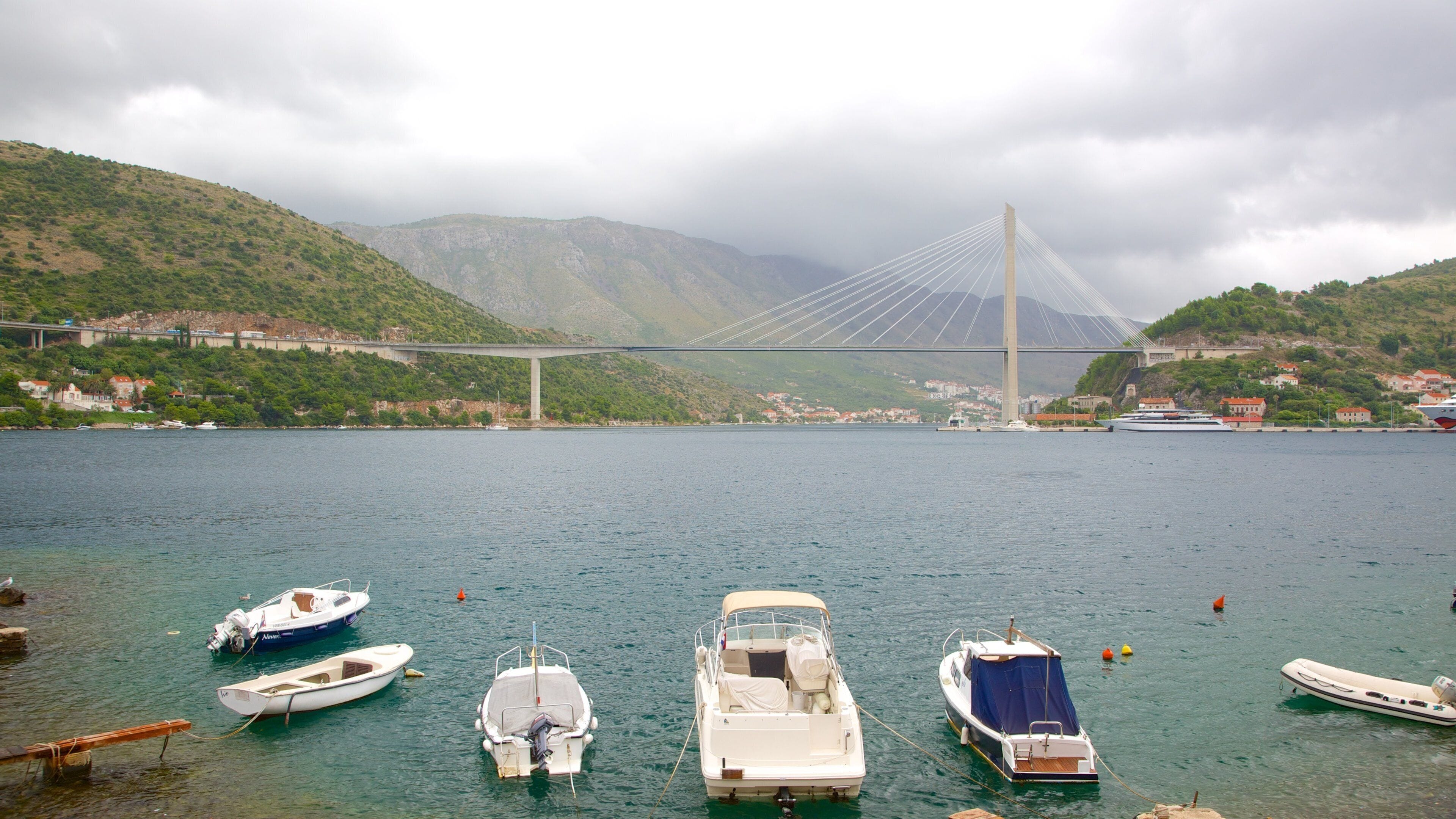 A bridge over a calm lake with boats docked in the foreground and a mountainous landscape in the background.