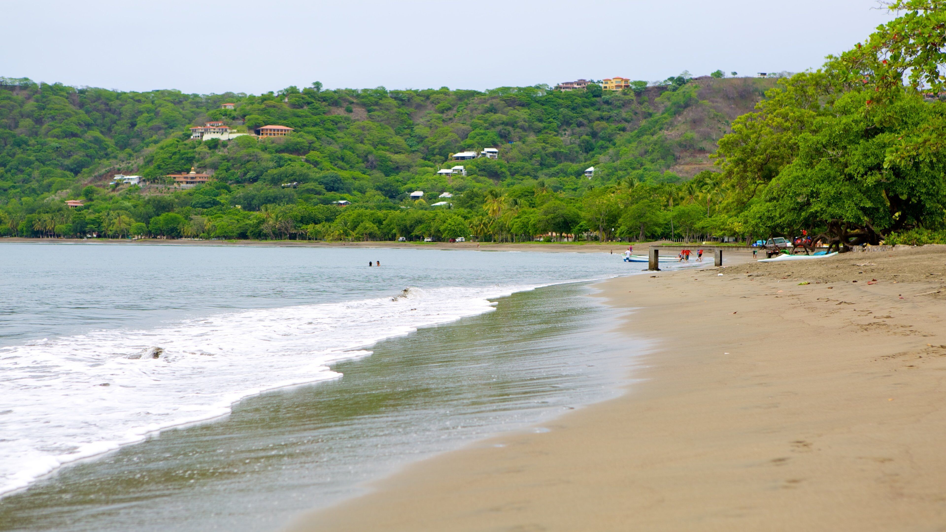 A beach with waves, a sandy shore, and houses on a hillside in the background.