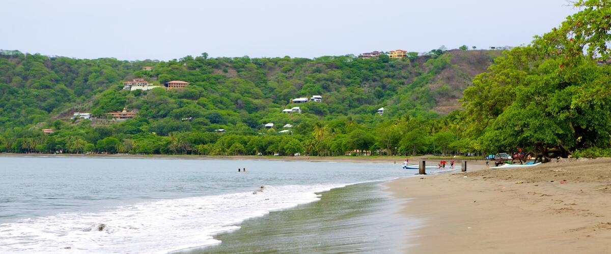 A beach with waves, a sandy shore, and houses on a hillside in the background.