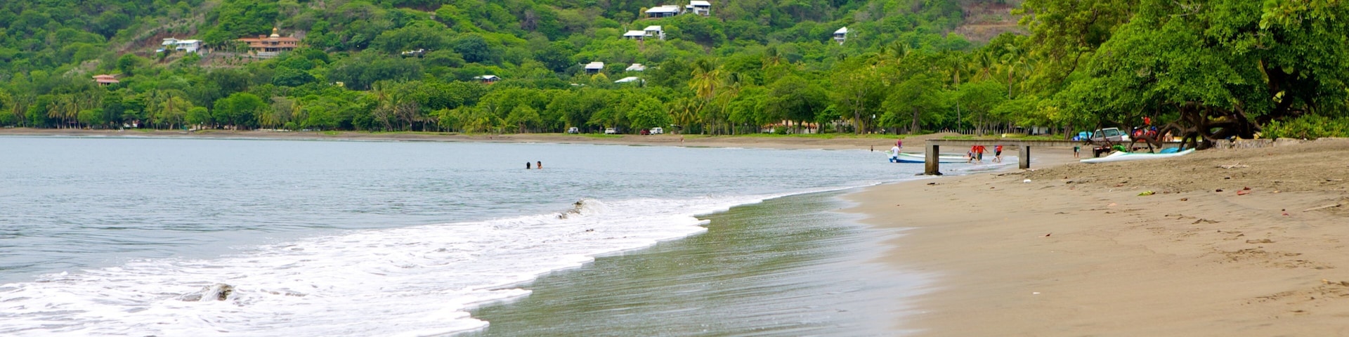 A beach with waves, a sandy shore, and houses on a hillside in the background.