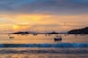 A sunset over a calm sea with multiple boats anchored, surrounded by a hilly landscape.