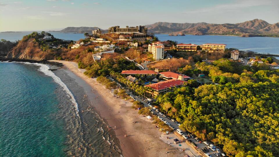 A coastal town with a beach, a large building complex, and mountains in the background.