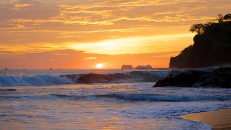 Ein Sonnenuntergang über einem Strand, mit brechenden Wellen und einer felsigen Klippe am rechten Rand.