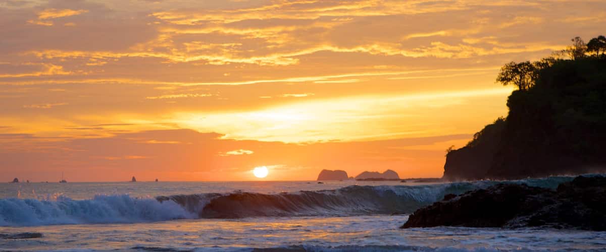 A sunset over a beach with waves crashing onto the shore and a rocky cliff on the right.