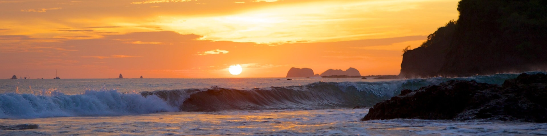 A sunset over a beach with waves crashing onto the shore and a rocky cliff on the right.