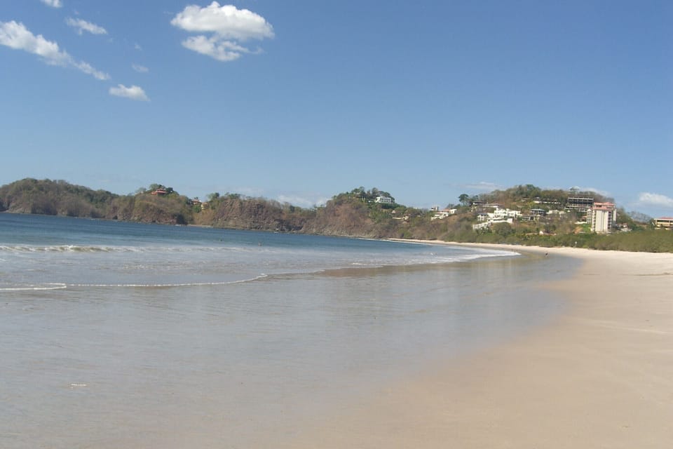 A beach with gentle waves, a clear sky, and buildings in the background.