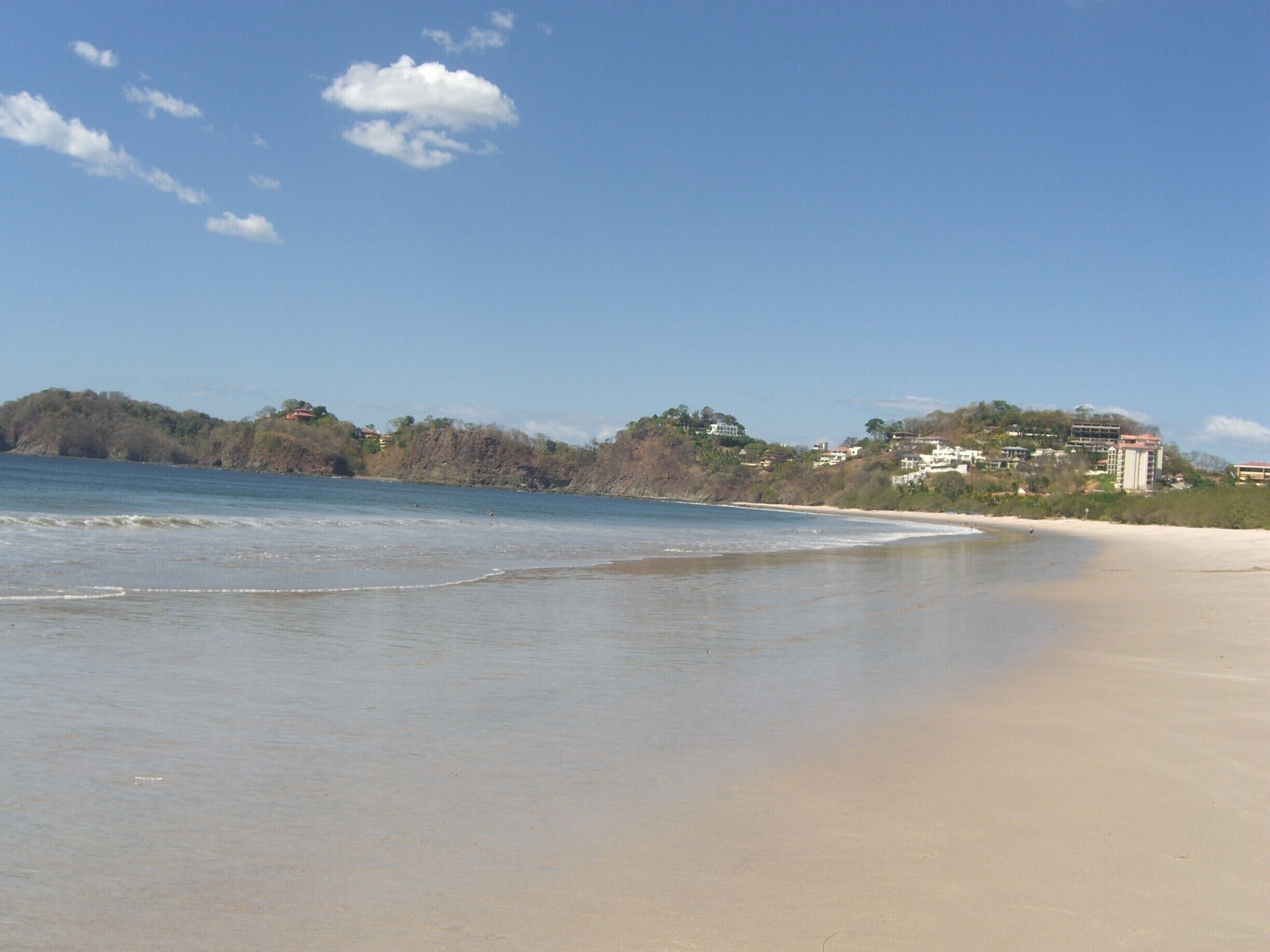 A beach with gentle waves, a clear sky, and buildings in the background.