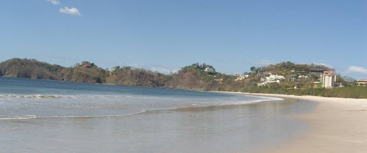 A beach with gentle waves, a clear sky, and buildings in the background.