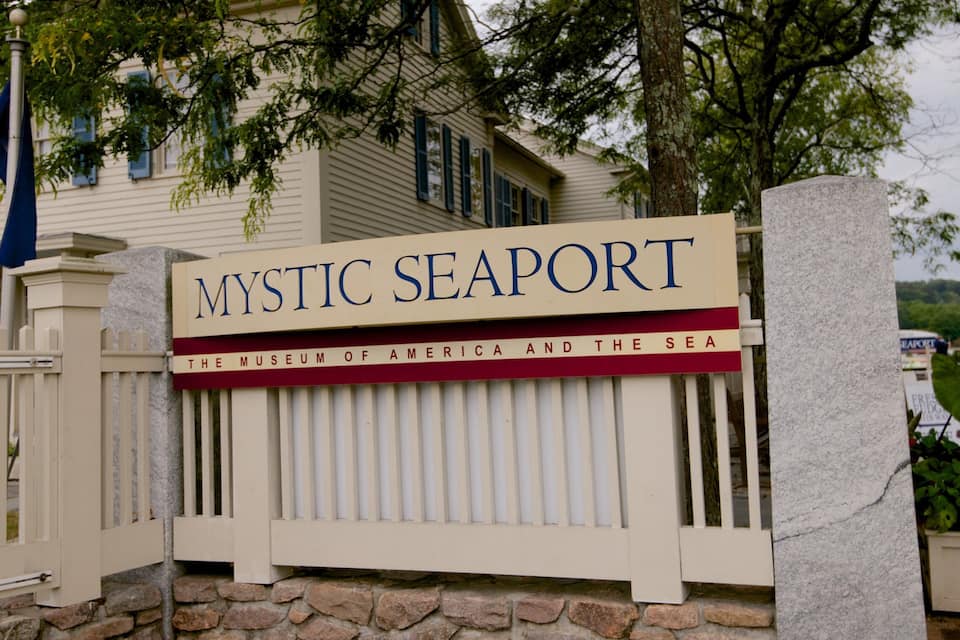 A sign for Mystic Seaport, the Museum of America and the Sea, with a stone wall and a wooden fence.