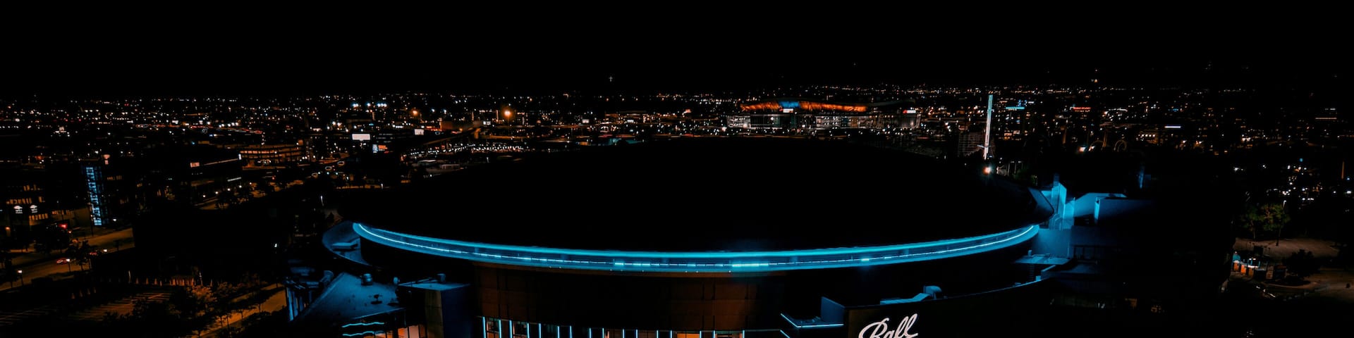 A brightly lit building with a circular design, surrounded by a parking area and a cityscape in the background.