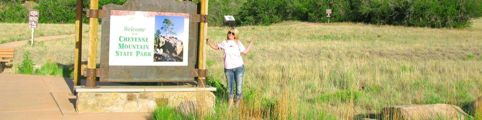 A wooden signpost with a welcome message to Cheyenne Mountain State Park, a mountainous backdrop, and a grassy field.