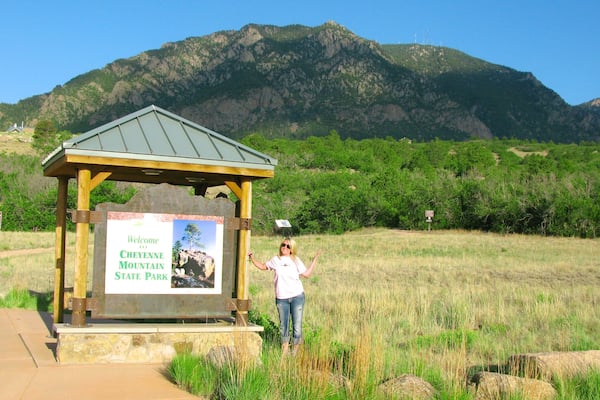 A wooden signpost with a welcome message to Cheyenne Mountain State Park, a mountainous backdrop, and a grassy field.