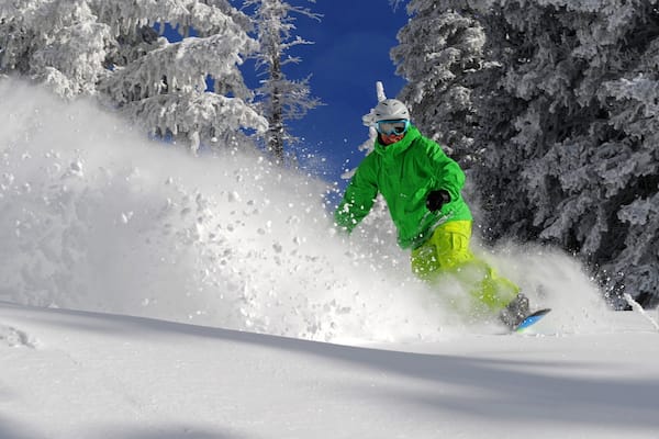 Uma pessoa pratica snowboard em uma encosta nevada, com árvores ao fundo.