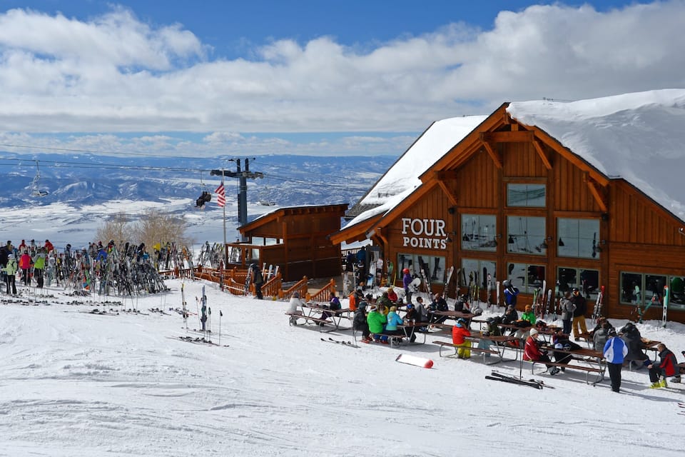 A wooden ski lodge with a sign reading "FOUR POINTS" amidst a snowy landscape with skiers and a clear blue sky.