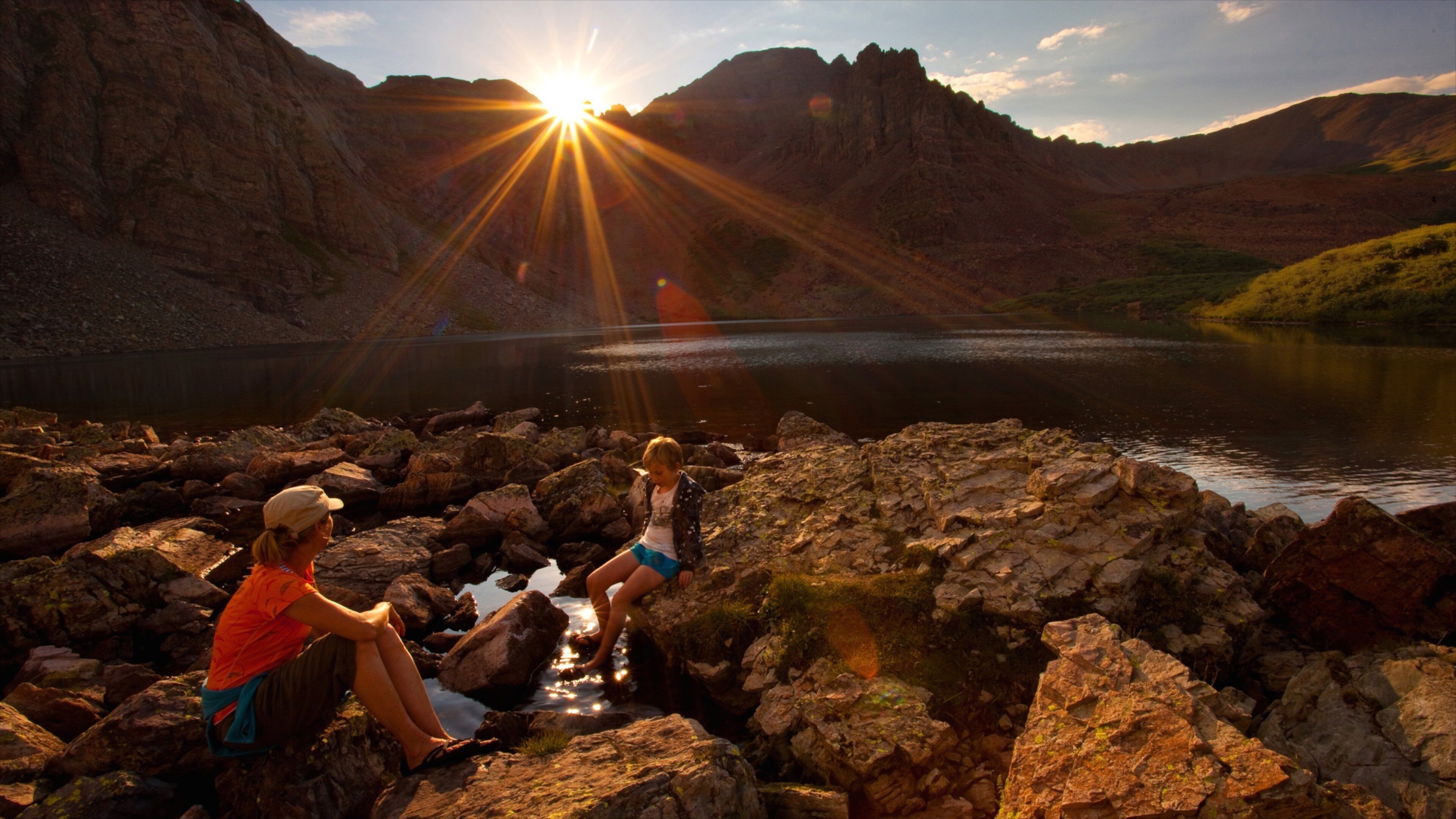 Two people sitting on rocks by a lake with mountains in the background and the sun shining.