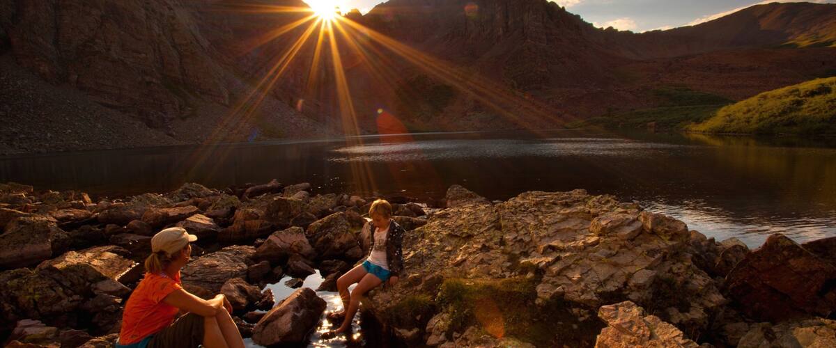 Two people sitting on rocks by a lake with mountains in the background and the sun shining.