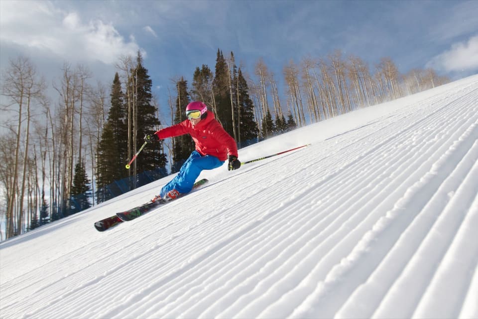 A skier in a red jacket and blue pants skiing down a snowy slope with trees in the background.
