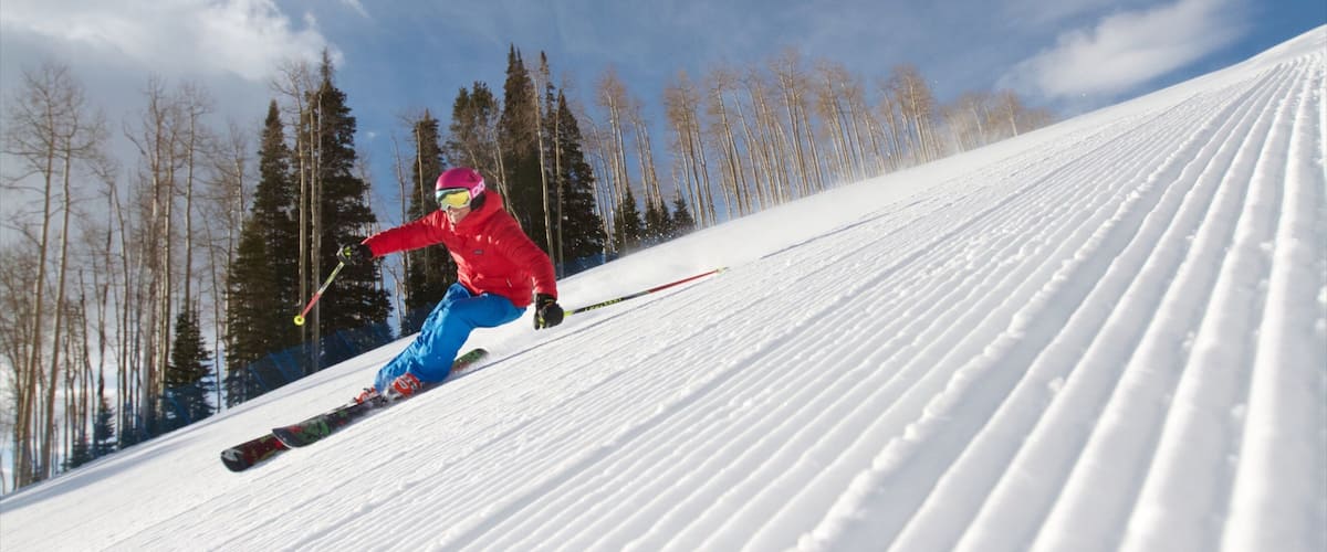 A skier in a red jacket and blue pants skiing down a snowy slope with trees in the background.