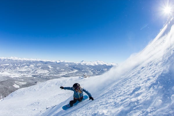 Ein Snowboarder befindet sich auf einer steilen, mit Schnee bedeckten Piste, mit blauem Himmel und in der Ferne schneebedeckten Bergen.