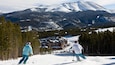 Des skieurs sur une pente enneigée, avec un chalet et des montagnes en arrière-plan.