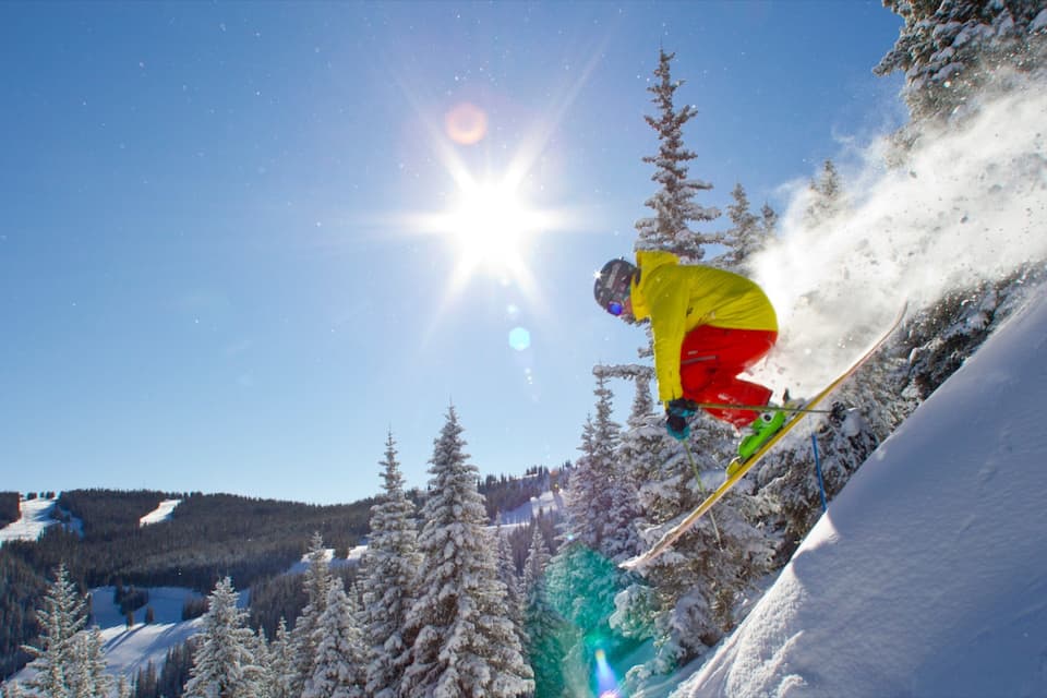 A skier in a yellow jacket and red pants performing a jump on a snowy slope with trees and a clear blue sky in the background.