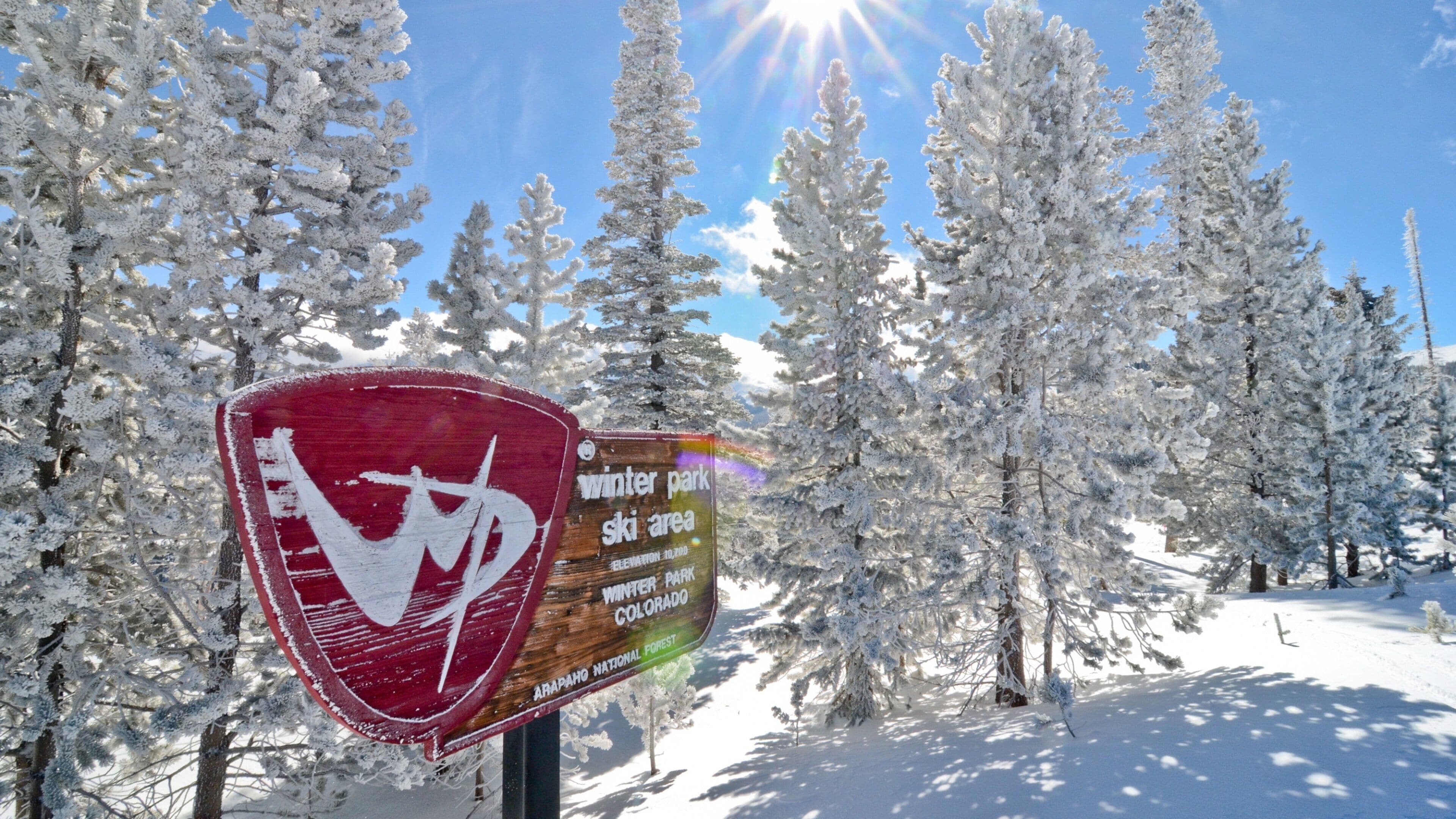 Cartel de una estación de esquí cubierta de nieve, con un símbolo de esquí, rodeado de árboles cargados de nieve y un cielo azul despejado.