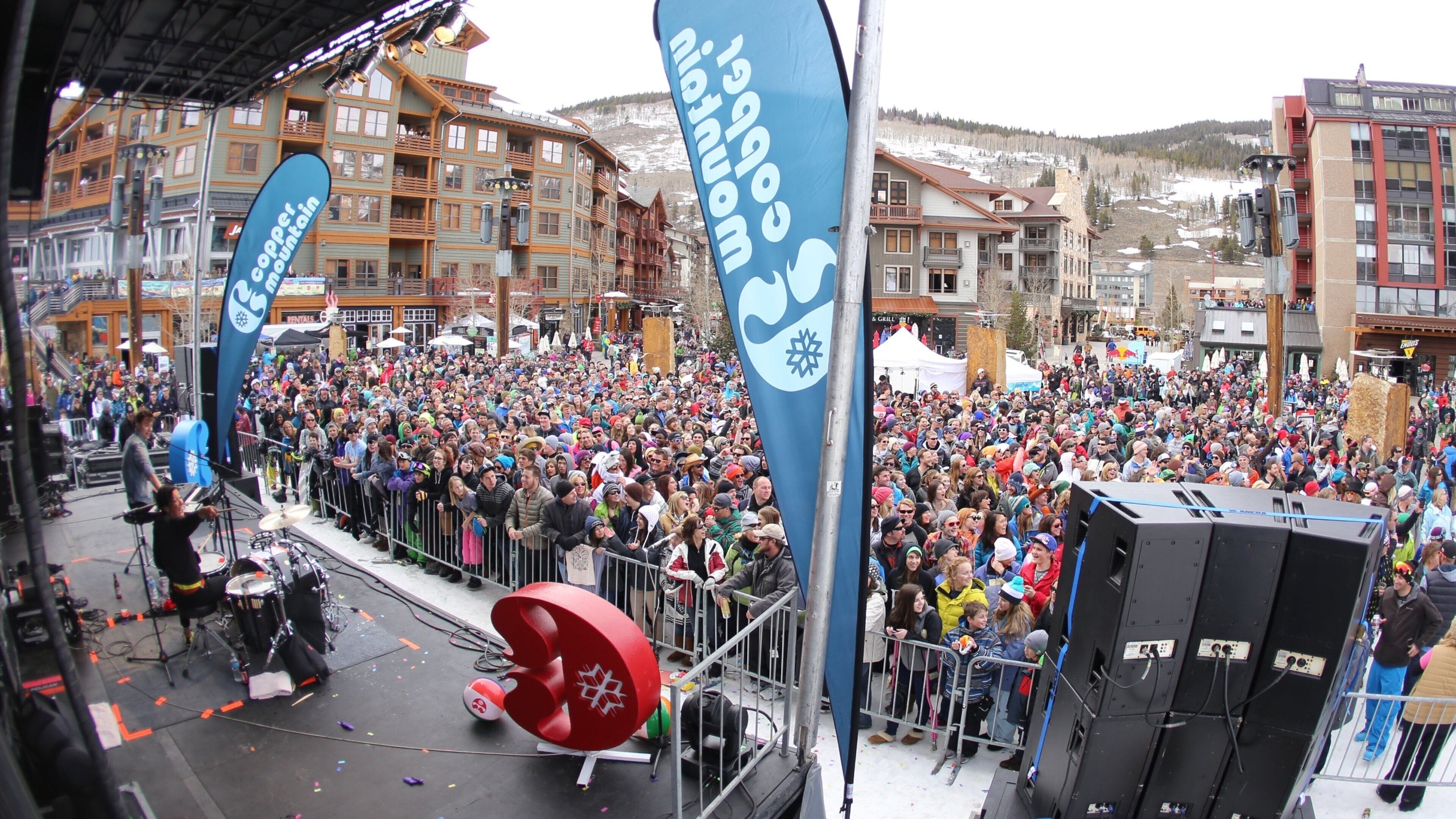 A crowded outdoor event with a stage, large audience, and buildings in the background.