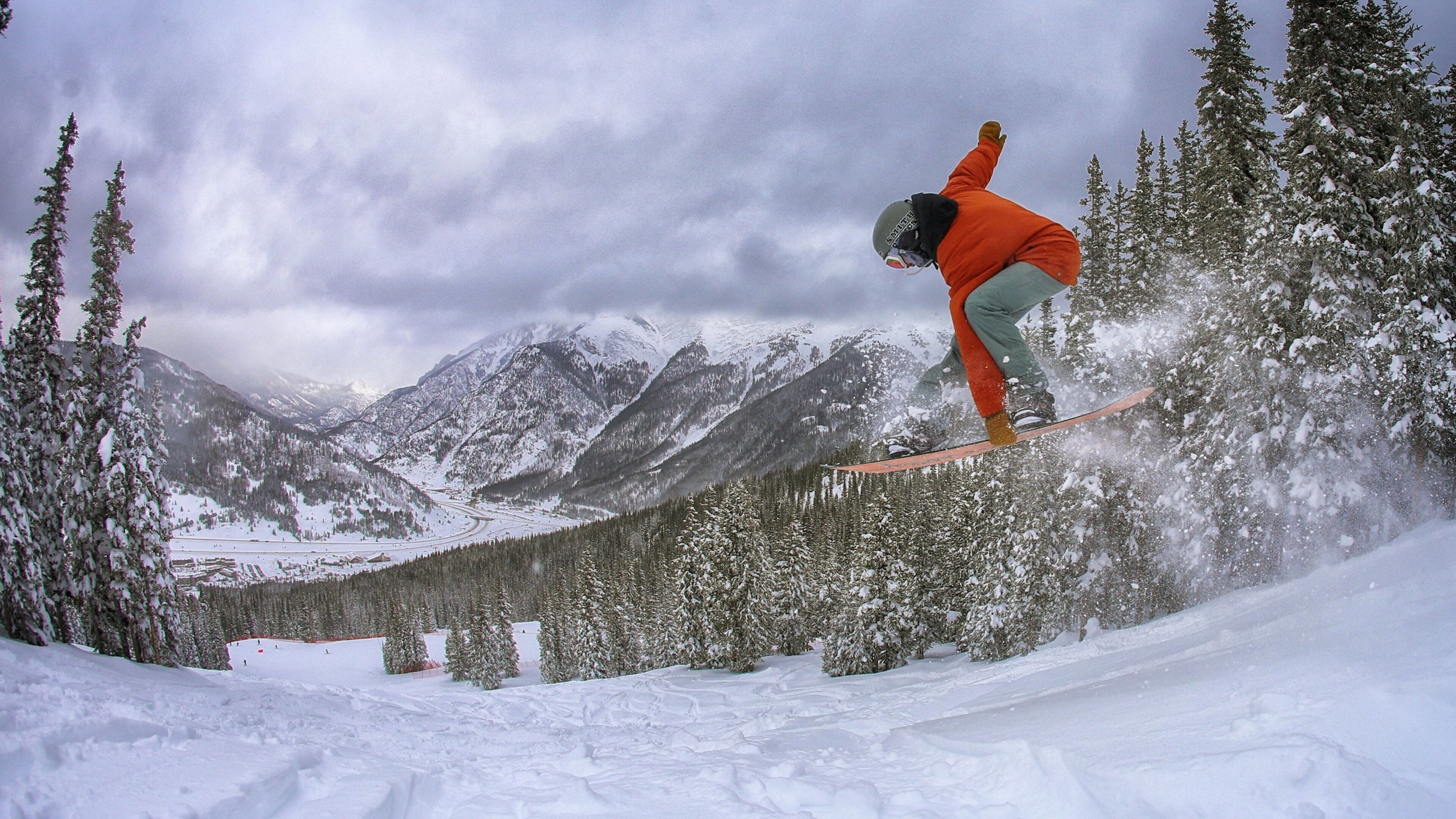 A snowboarder in mid-air performing a jump in a snowy mountainous area with evergreen trees and snow-covered peaks in the background.
