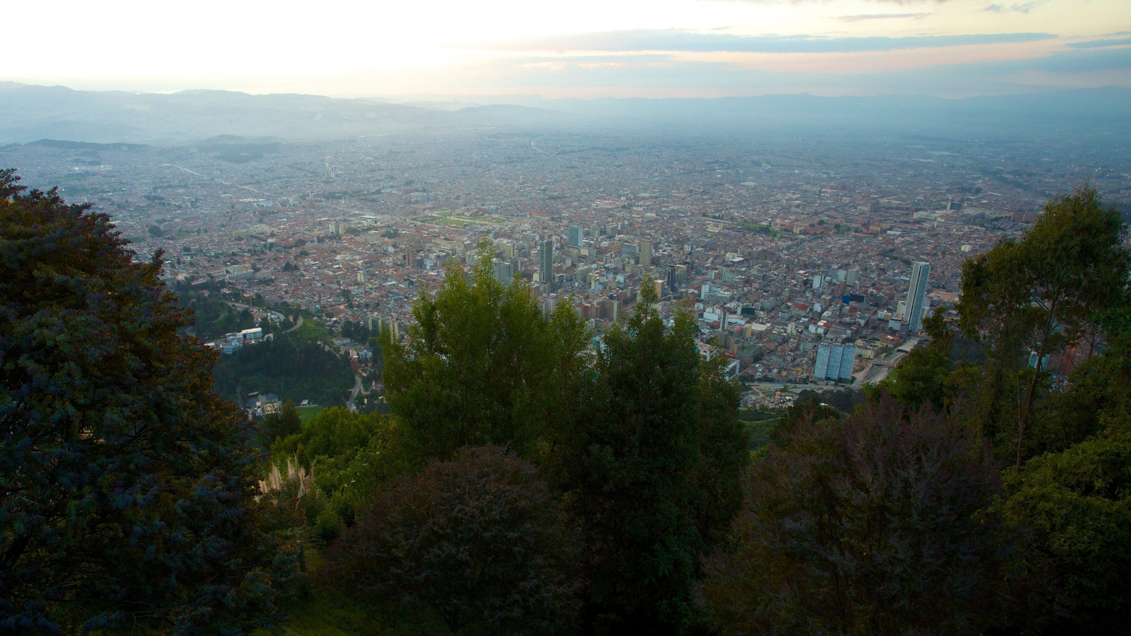 Vista panorâmica de uma cidade a partir de um ponto alto, com edifícios urbanos densos e uma cadeia de montanhas ao longe.