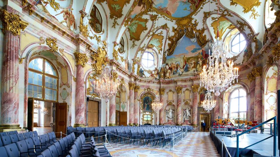 Un salón suntuoso con detalles dorados ornamentados, grandes candelabros y filas de sillas.