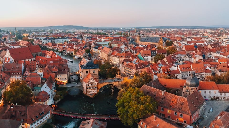 Une vue panoramique d’une ville historique, avec ses bâtiments aux toits rouges, une rivière et un pont imposant.