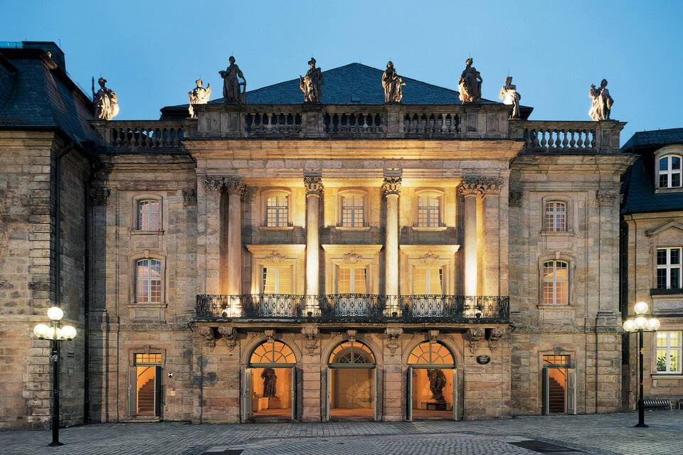 Un edificio histórico con arquitectura clásica, que presenta un balcón con estatuas y una entrada imponente.