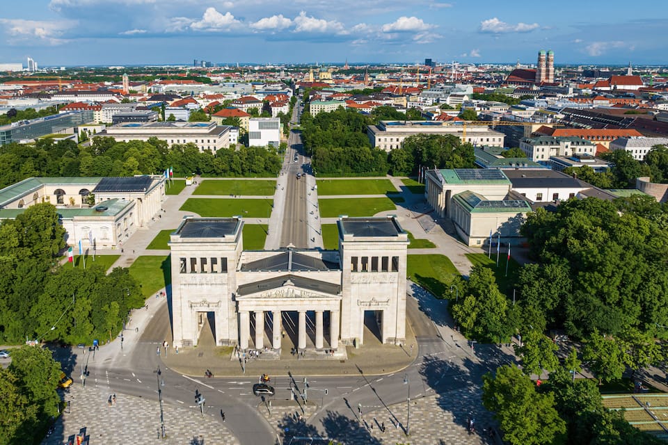 Un grand bâtiment historique doté d’une tour centrale, entouré d’espaces verts et bordé par une rue animée.